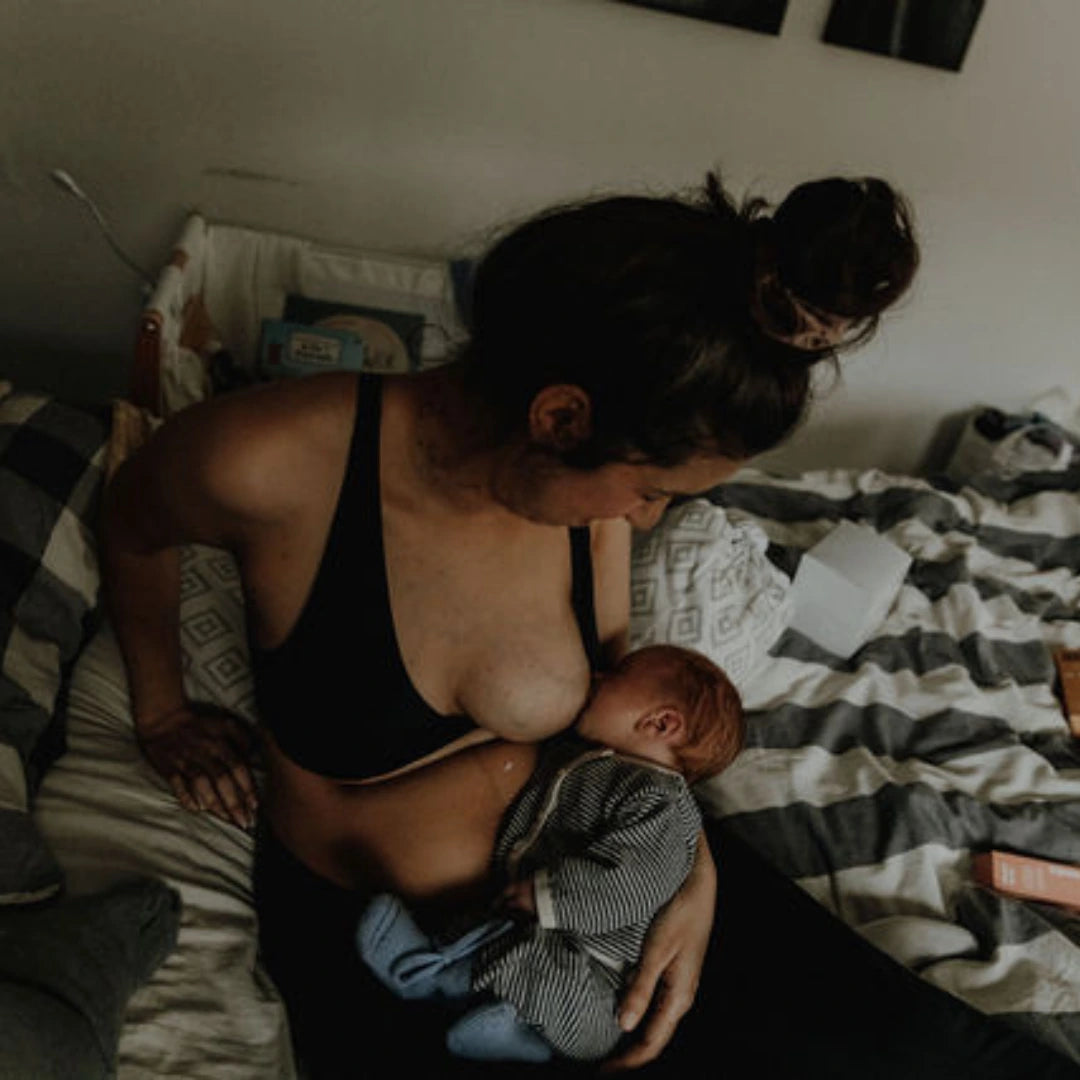 Frau sitzt auf dem Bett und stillt ihr Baby, im Hintergrund stehen Stilleinlagen von the weeks