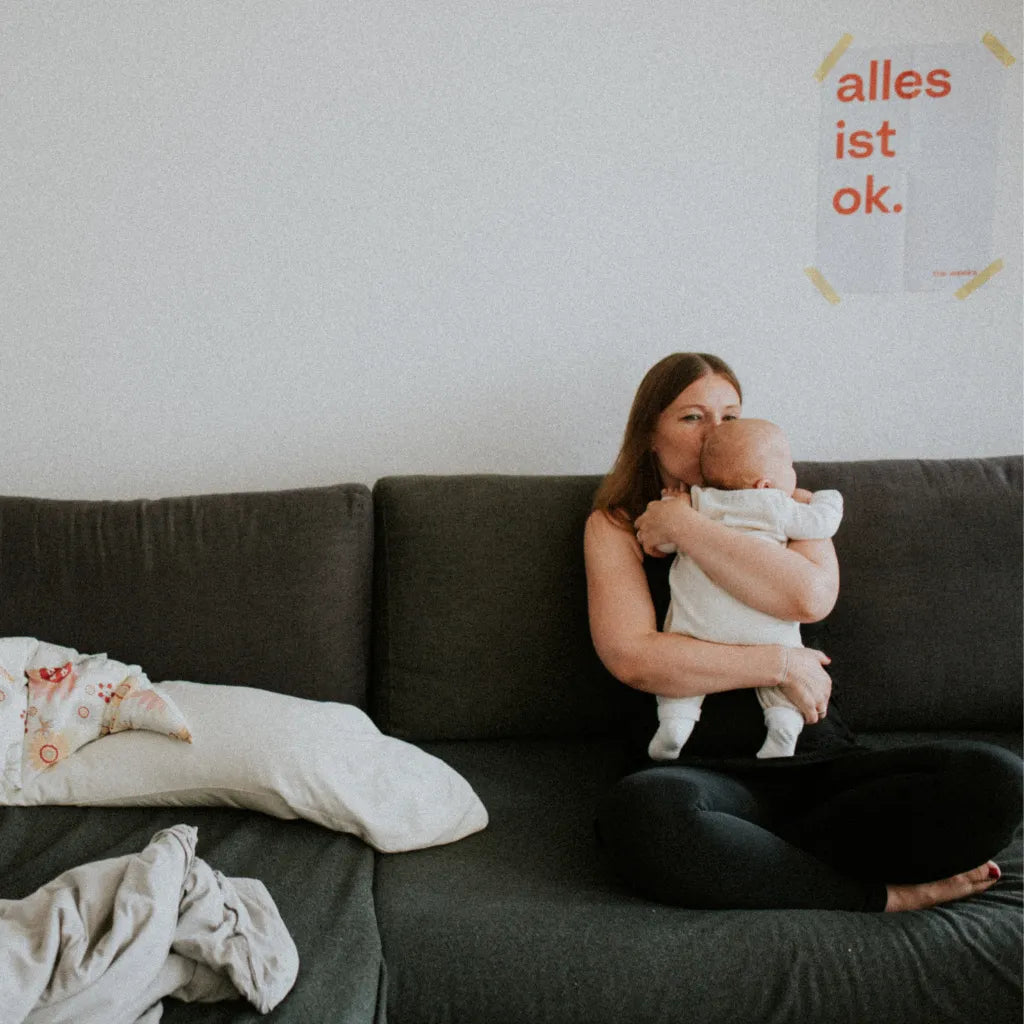 Frau sitzt auf dem Sofa mit einem kleinen Baby, an der Wand hängt das Wochenbett-Poster von the weeks