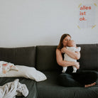 Frau sitzt auf dem Sofa mit einem kleinen Baby, an der Wand hängt das Wochenbett-Poster von the weeks