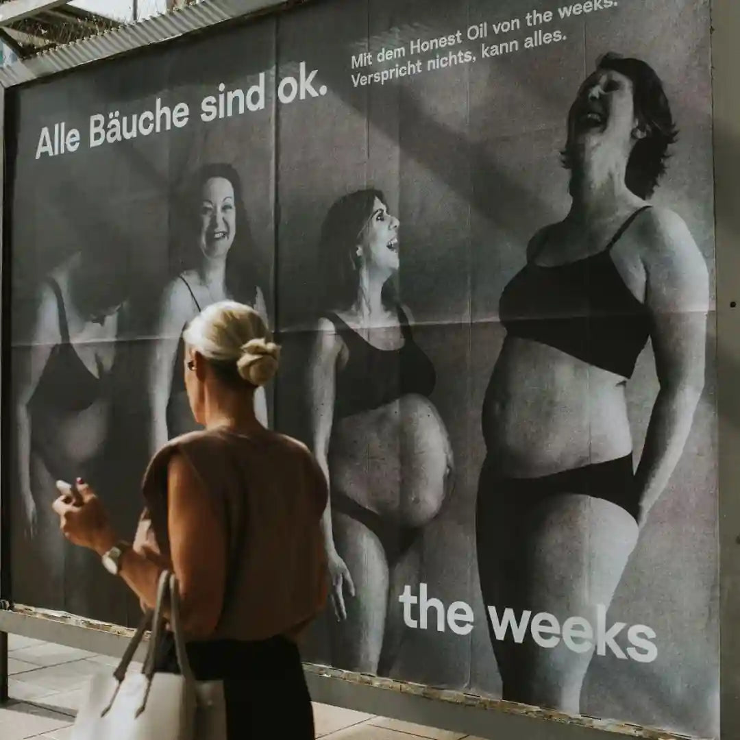 Frau geht an einem Plakat vorbei. Auf dem Plakat sind vier schwangere Fraun zu sehen, die Überschrift lautet "Alle Bäuche sind ok." Der Subtitle lautet: "Mit dem Honest Oil von the weeks. Verspricht nichts, kann alles.