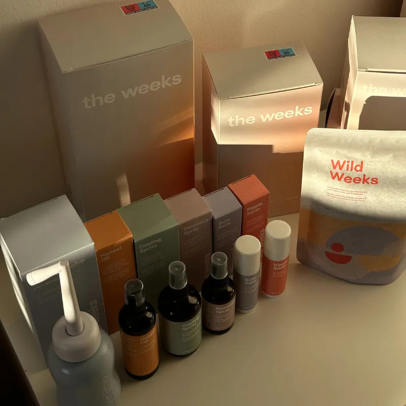 Produkte der WHOLE Wochenbettbox von the weeks stehen aufgereiht auf einem Tisch: Podusche, Hinest Oil, Cooling Spritz, Recovery Spray, Bum Balm, Nippel Saver, Tee Wild Weeks, Wochenbetteinlagen, Wochenbettbinden