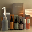 Produkte der WHOLE Wochenbettbox von the weeks stehen aufgereiht auf einem Tisch: Podusche, Hinest Oil, Cooling Spritz, Recovery Spray, Bum Balm, Nippel Saver, Tee Wild Weeks, Wochenbetteinlagen, Wochenbettbinden